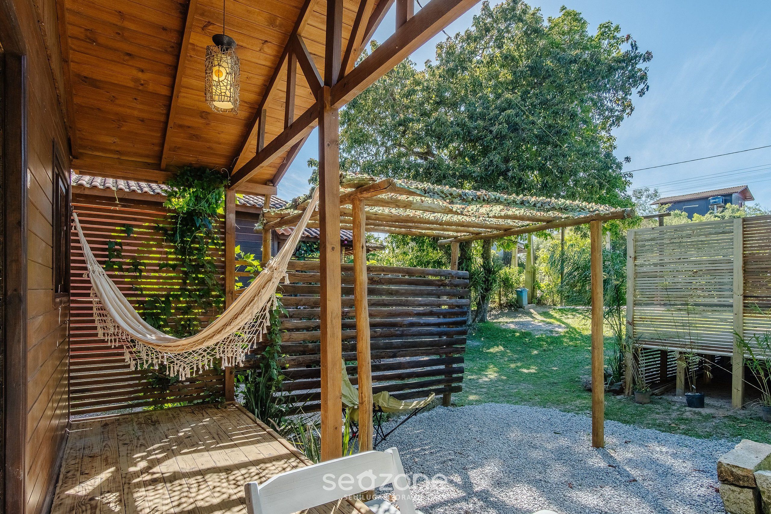 Una pérgola de madera con una hamaca, una terraza de madera y un jardín con valla y árboles.