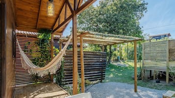 Una pérgola de madera con una hamaca, una terraza de madera y un jardín con valla y árboles.