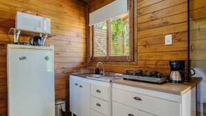 Cocina compacta con paredes de madera, un refrigerador blanco, estufa, cafetera y ventana con vista a la vegetación.