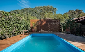 A rectangular swimming pool with a wooden deck surrounding it, a wooden fence, and lush greenery in the background. - Complete Chalet in Imbituba Rel0001 (Imbituba)