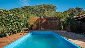 A rectangular swimming pool with a wooden deck surrounding it, a wooden fence, and lush greenery in the background.