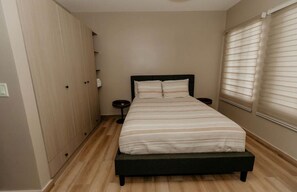 Ein Schlafzimmer mit einem Bett, einem Kleiderschrank, einem Nachttisch und einem Fenster mit Jalousien.
