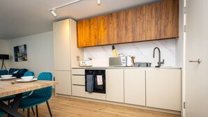 A modern kitchen with white cabinetry, a wooden upper shelf, and a dining area with a wooden table and teal chairs. - Birmingham City Retreat No 126 (Birmingham)