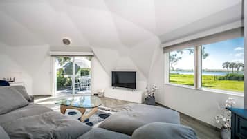 A modern living room with a glass coffee table, grey sofas, a flat-screen TV, and a large window with a view of a green landscape.