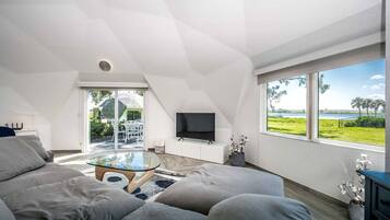 Een moderne woonkamer met een glazen salontafel, grijze banken, een flatscreen televisie en een groot raam met uitzicht op een groen landschap.