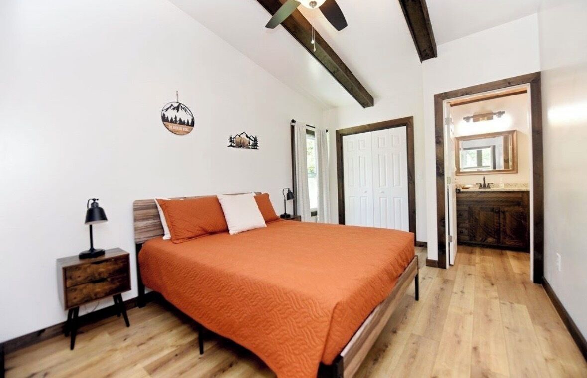 A bedroom with a bed, a nightstand, a wooden floor, a ceiling fan, and a door leading to a bathroom.