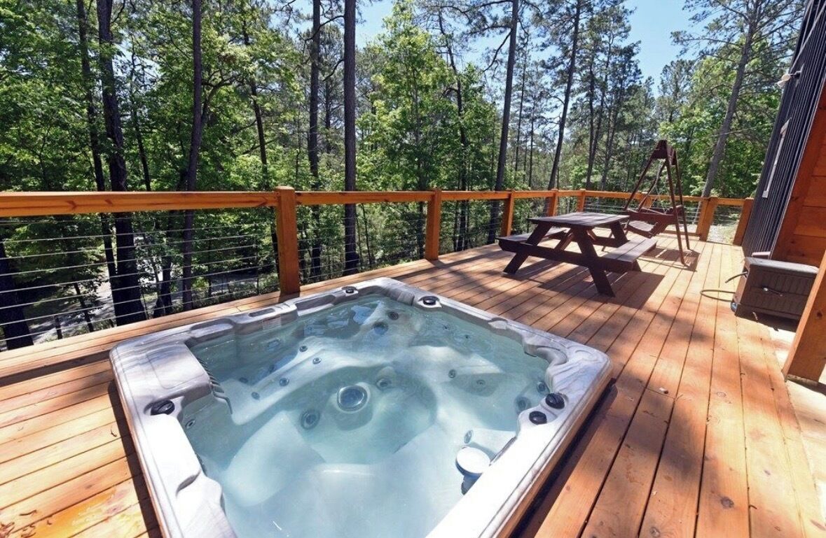 Un bain à remous sur une terrasse en bois, avec une table de pique-nique et une balançoire en arrière-plan, le tout entouré d’arbres.