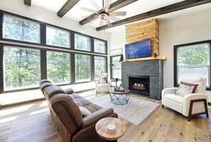 A spacious living room with a fireplace, large windows, and a ceiling fan. - Mountain Melody- Cozy 3-bedroom cabin with AC in Ellijay- Hot Tub + EV Charger (Ellijay)