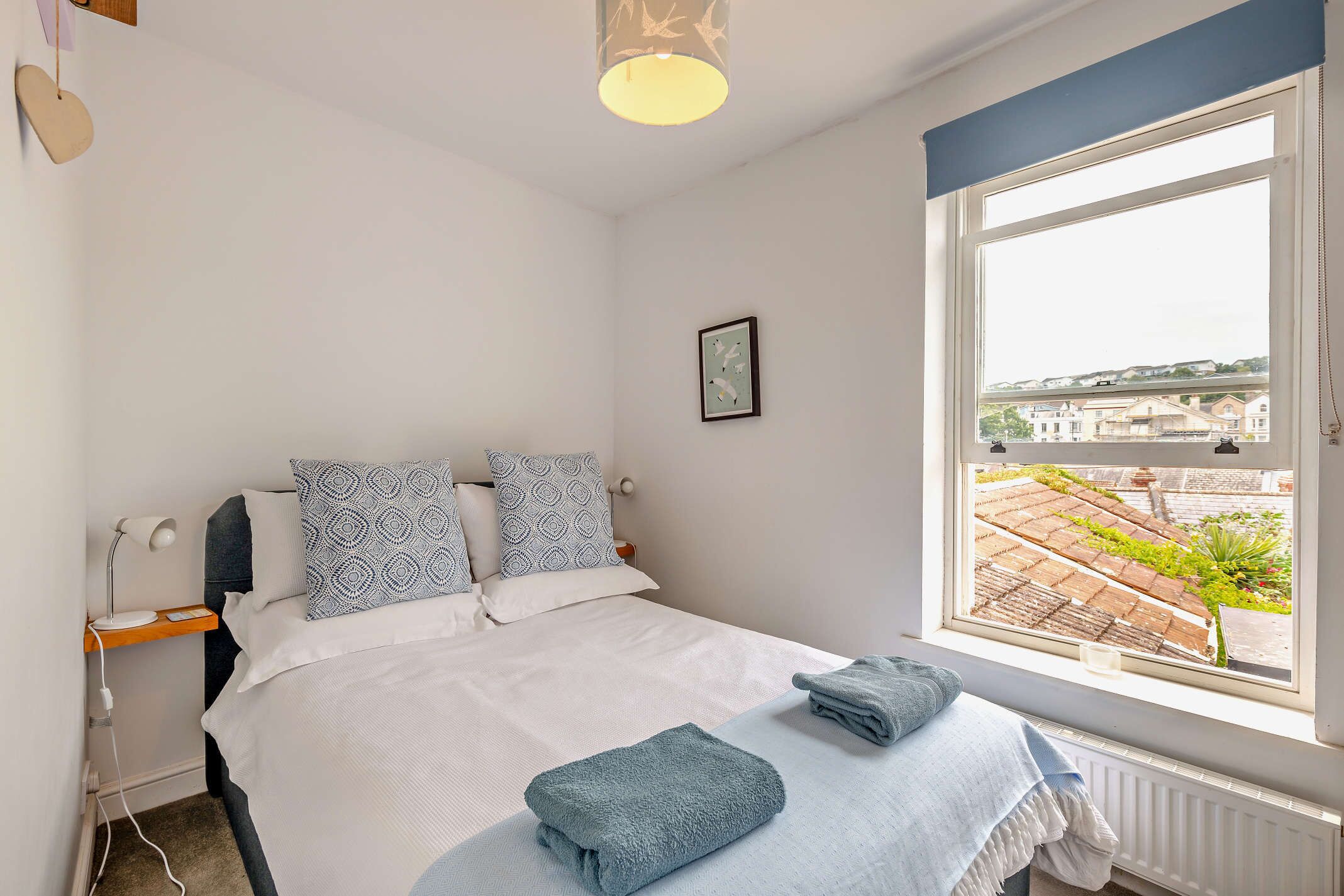 A bedroom with a bed, pillows, a bedside table with a lamp, a window with a view of buildings, and a framed picture on the wall.