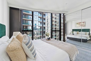 Ein modernes Schlafzimmer mit einem großen Bett, einem kleineren Bett und Blick auf einen Hochhaus durch raumhohe Fenster.