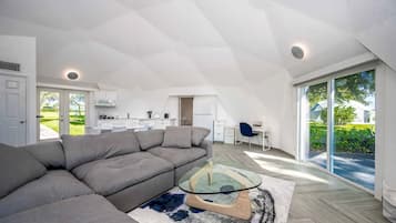 Un moderno loft con un ampio divano angolare grigio, un tavolino in vetro e vista su un rigoglioso giardino attraverso ampie porte scorrevoli in vetro.