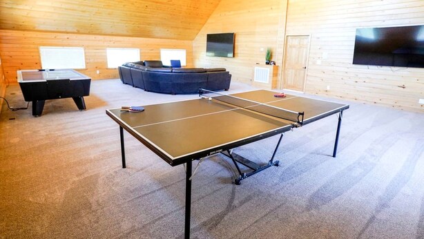 Et bordtennisbord i et rum med træloft og vægge, en blå sofa og et fladskærms-tv monteret på væggen.