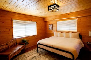 A wooden cabin bedroom with a bed, two windows with blinds, a bench, and a hanging light fixture. - Enormous Cabin with Game Room Perfect for Large Groups in Orderville, Utah (Orderville)