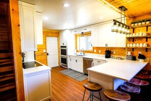 A modern kitchen with white cabinets, wooden flooring, and a central island with stools. - Enormous Cabin with Game Room Perfect for Large Groups in Orderville, Utah (Orderville)