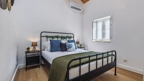 A bedroom with a bed, bedside tables, an air conditioning unit, a window, and a wooden floor. - Casa Maria by LovelyStay (Trafaria)