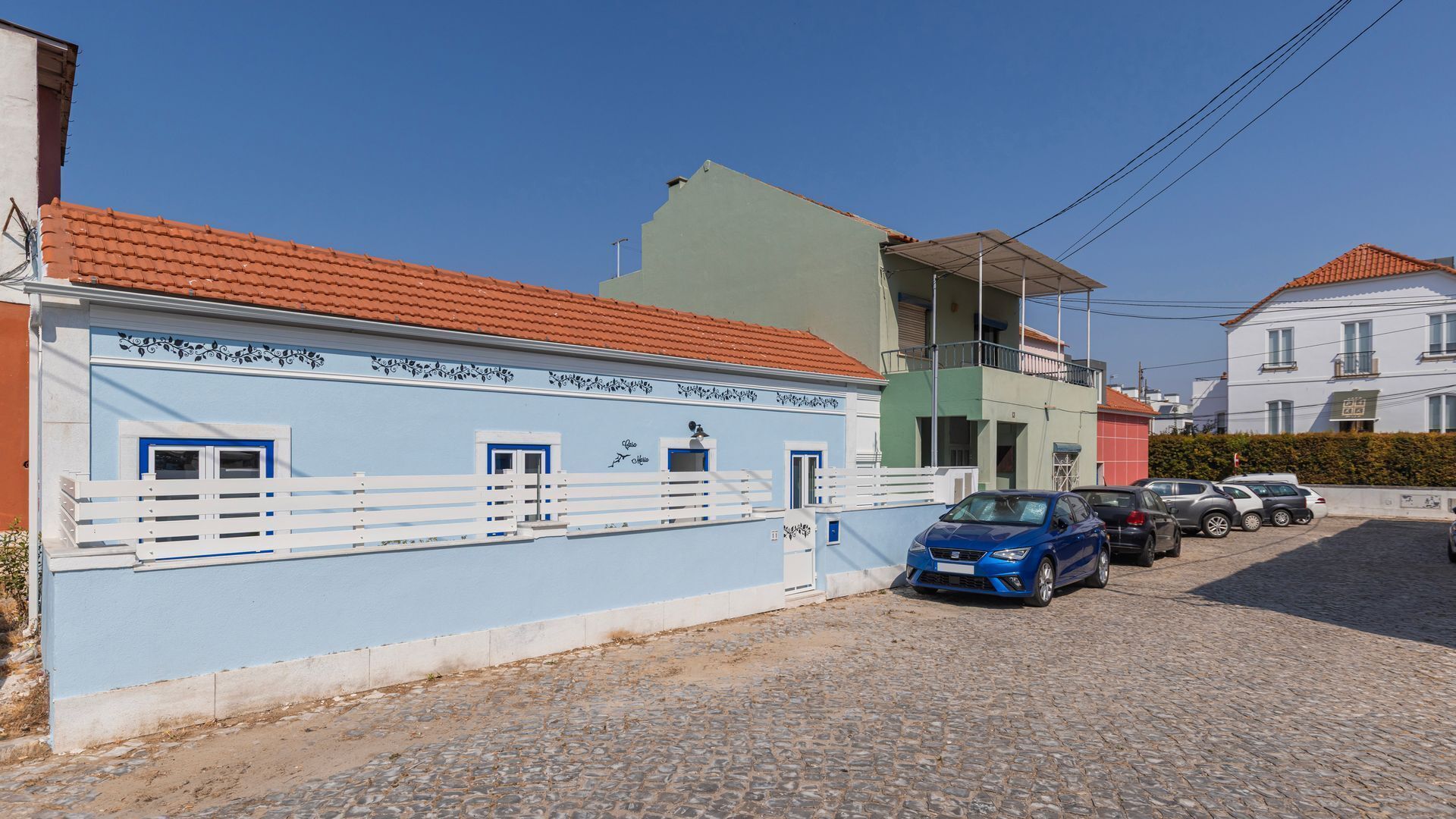 Une rue pavée de pierres, un immeuble bleu avec des balcons blancs et des voitures garées.
