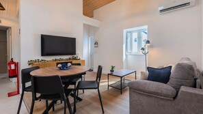 A modern living room with a flat-screen TV, a dining area with a wooden table and chairs, a sofa, and a window with a view. - Casa Maria by LovelyStay (Trafaria)