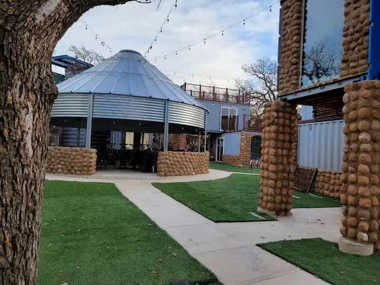 Un edificio con techo abovedado, rodeado de una zona pavimentada con parterres de césped y pilares de piedra.