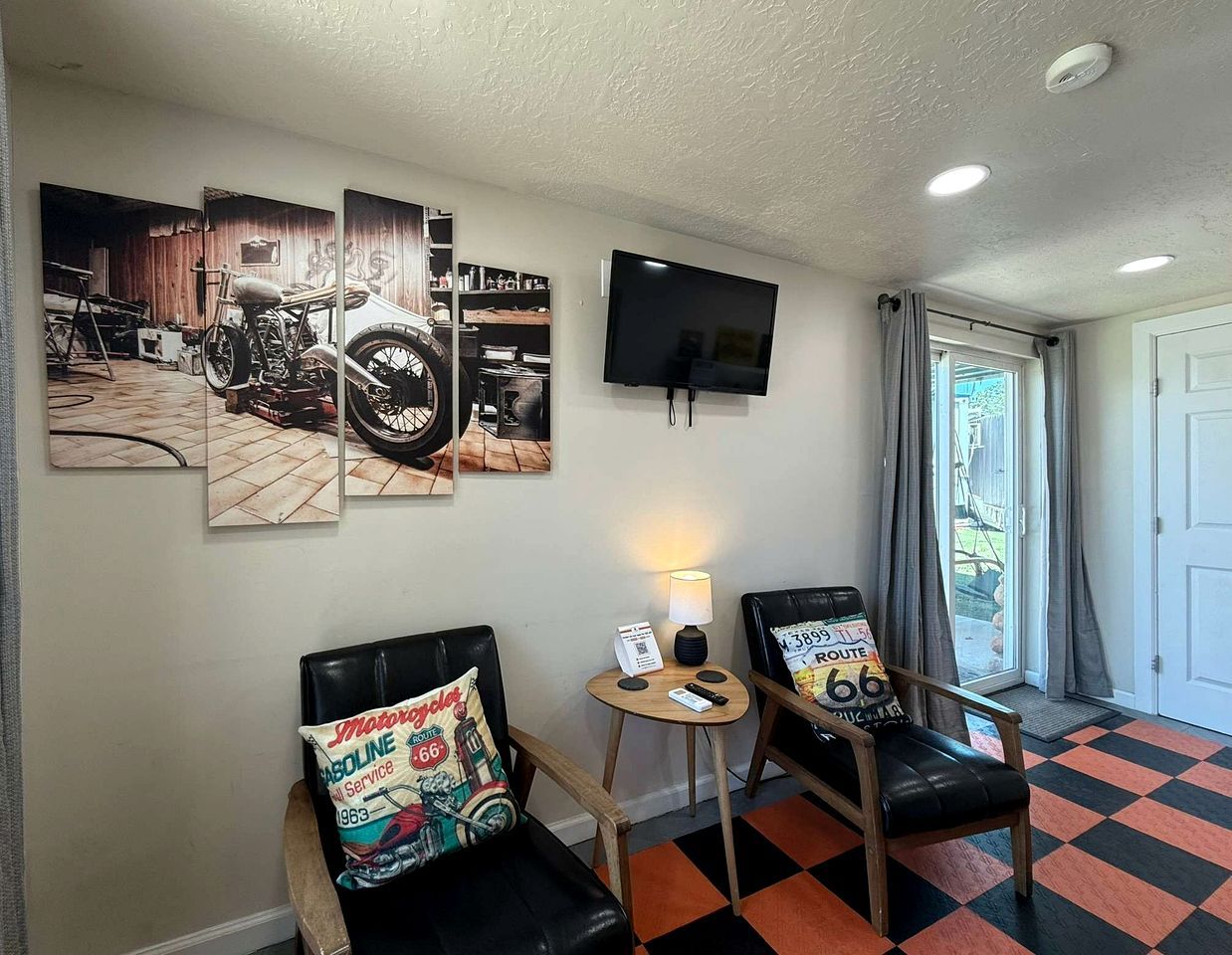 Een kamer met twee zwarte leren stoelen, een bijzettafeltje met een lamp, een flatscreen-televisie aan de muur en een geruit vloerkleed.