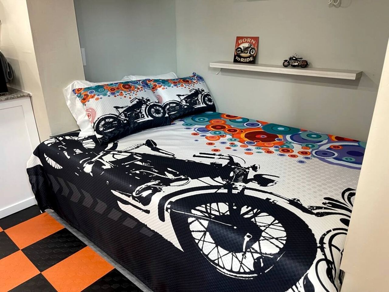A bed with a motorcycle-themed bedspread, a pillow with a similar design, and a small shelf with a motorcycle model and a book titled 'Born to Ride'.