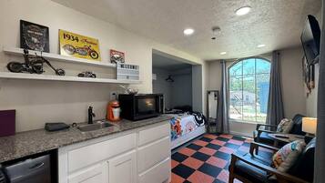 A compact room with a kitchenette, a bed, a sofa, and a window with curtains.