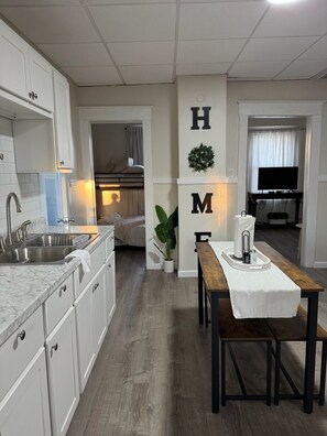 A compact kitchen with white cabinets, a wooden dining table, and a visible TV in the adjacent living area. - Cozy 2DR Apartment — Near Downtown Schenectady & Union College (Schenectady)