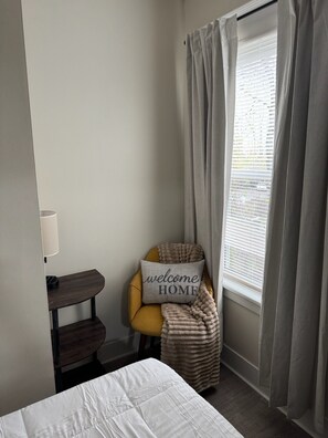 Ein Schlafzimmer mit Bett, einem Sessel mit Kopfkissen, einem Nachttisch und einem Fenster mit Vorhängen.