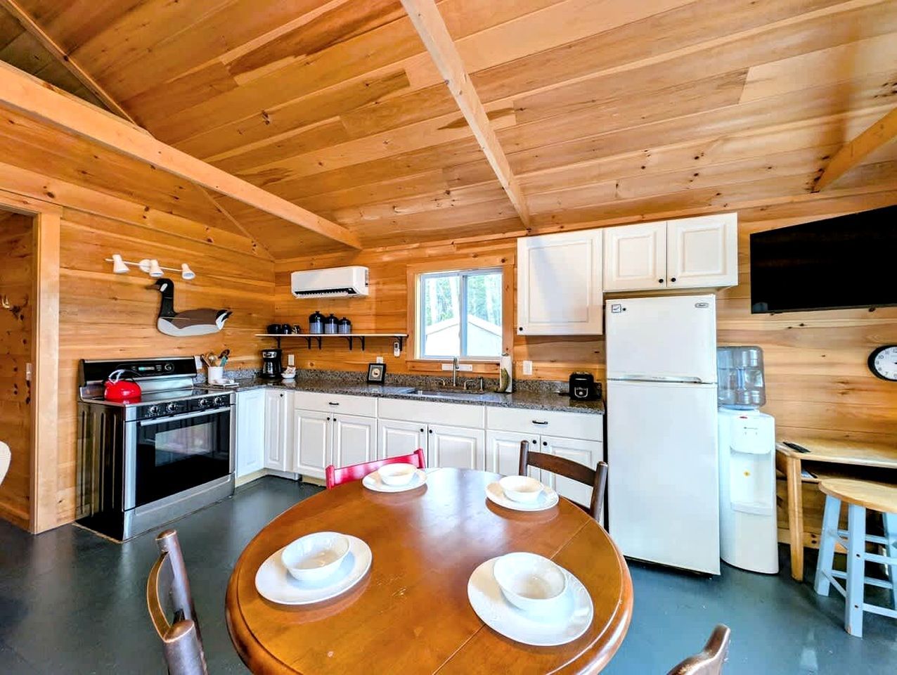 A wooden interior with a kitchen featuring white cabinets, a stove, and a refrigerator. There is a dining table with chairs and plates.