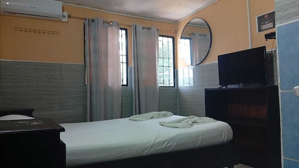 A single bed with a white mattress, a flat-screen TV on a stand, and a window with curtains.
