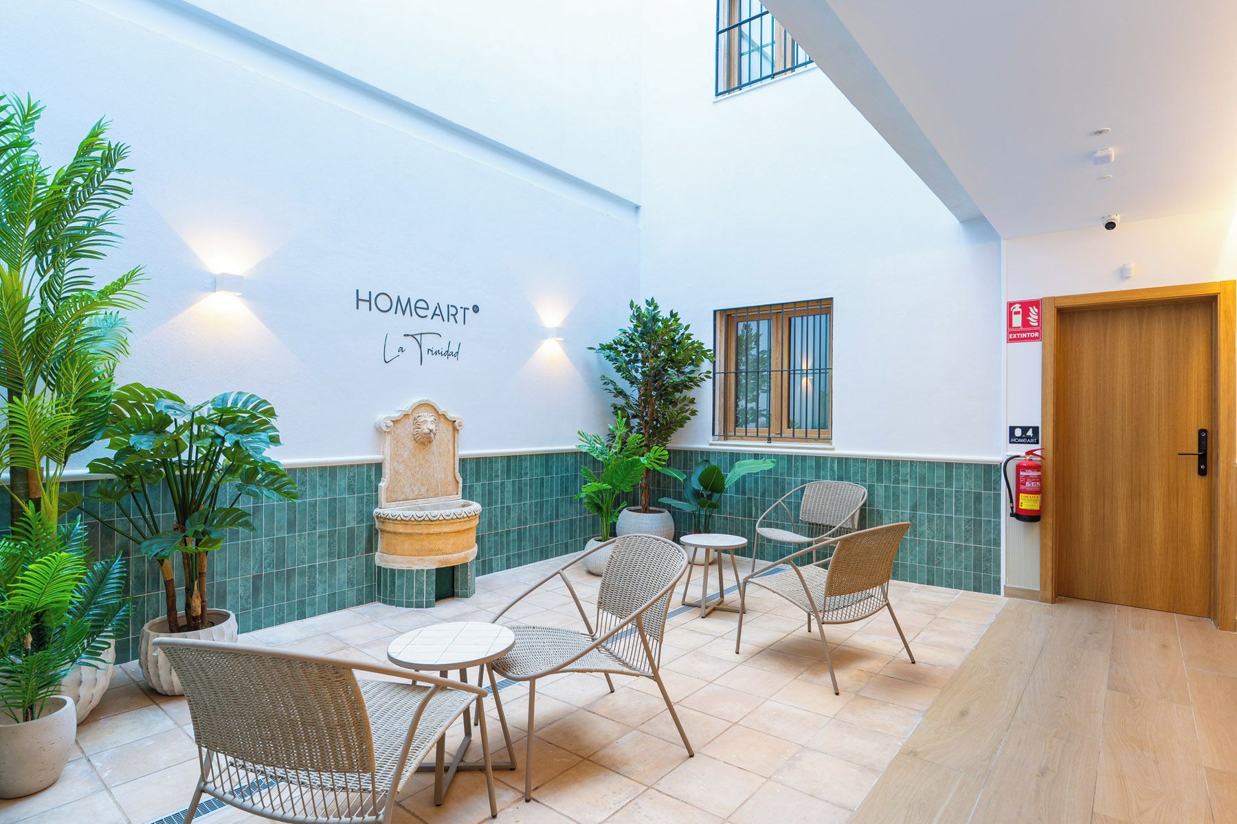 A hotel interior with a lounge area featuring wicker chairs, a stone fountain, and potted plants.