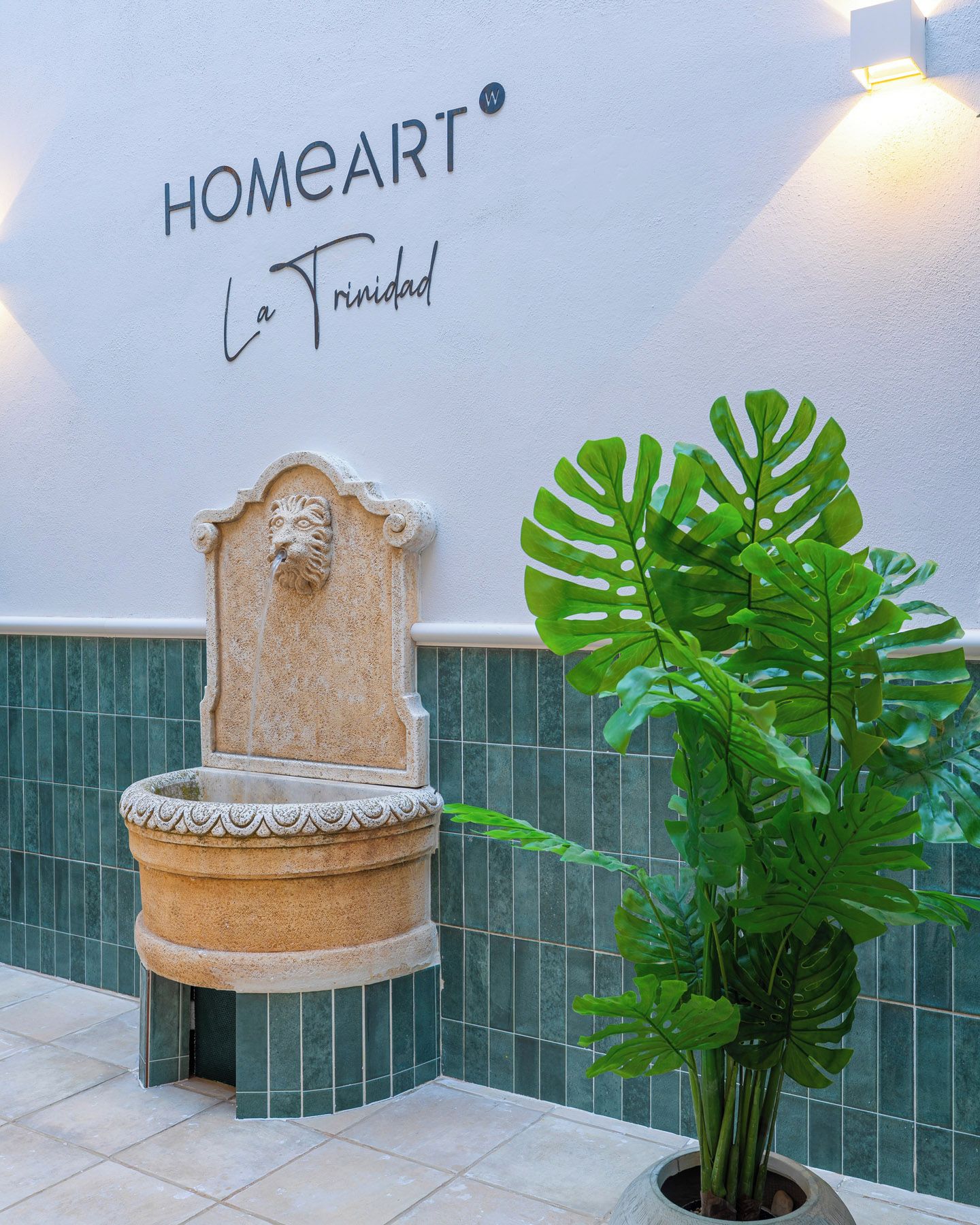Un mur orné d’une enseigne « HOMEART La Trinidad », une fontaine en pierre et une grande plante Monstera.