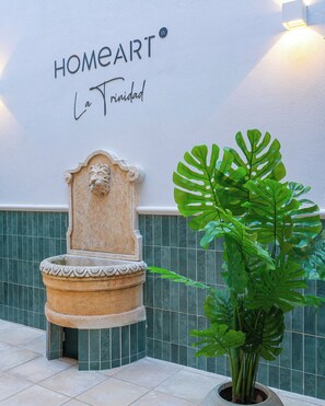 Eine Wand mit der Beschilderung 'HOMEART La Trinidad', einem Steinfountain und einer großen Monstera-Pflanze.