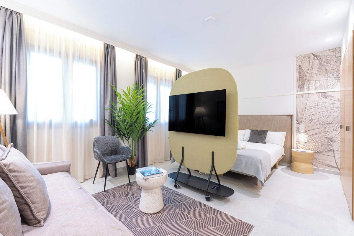 Ein modernes Hotelzimmer mit einer Couch, einem Sessel, einem an der Wand montierten Fernseher, einem Bett, einer Pflanze und einem kleinen Tisch.