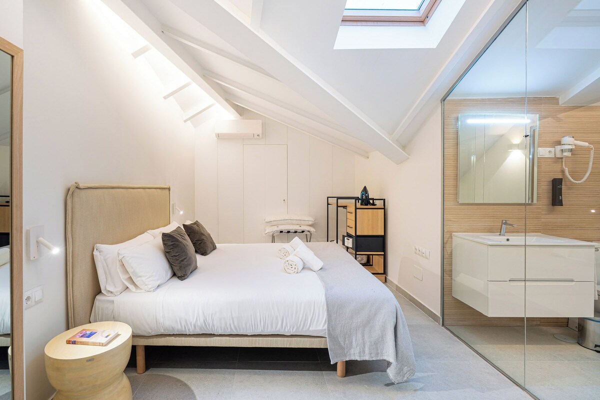Ein modernes Schlafzimmer mit Dachfenster, einem Bett mit Kissen, einem Nachttisch und einem Badezimmer mit Spiegel und Waschbecken.