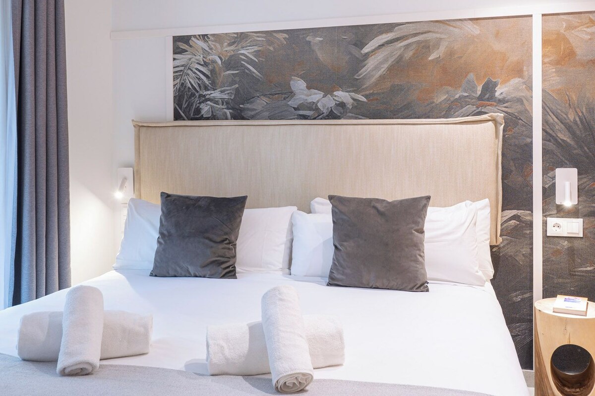 Ein Hotelzimmer mit einem großen Bett, einem Kopfteil mit floralem Wandbild und einem Nachttisch mit einem Buch und einem Lautsprecher.