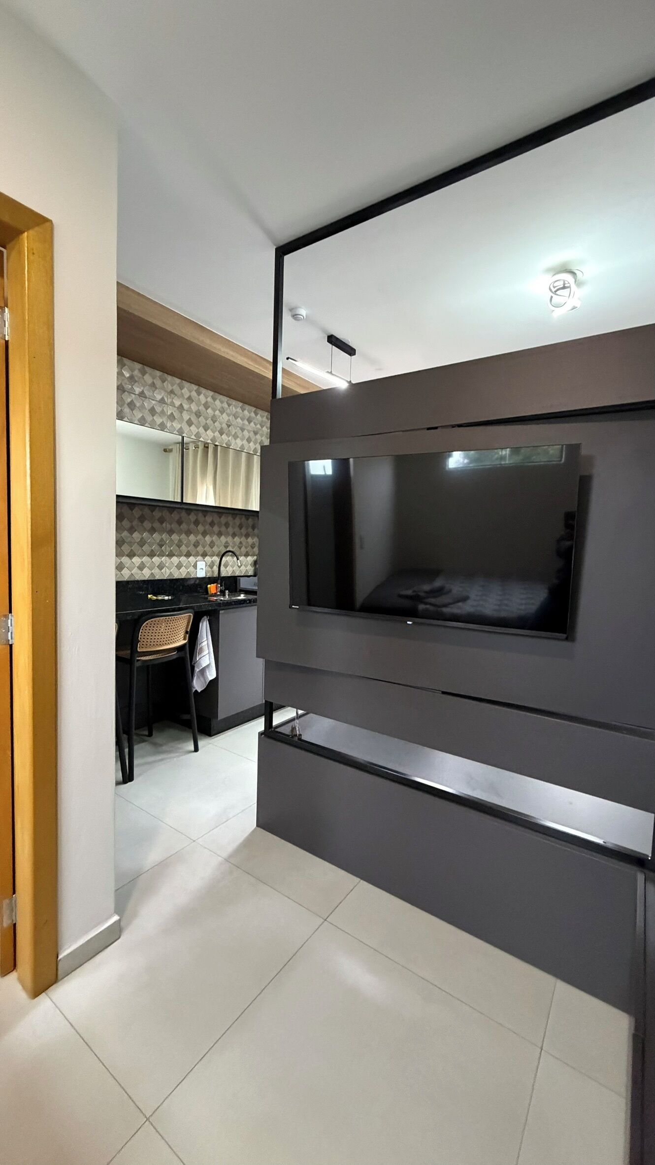 Una habitación moderna con un televisor de pantalla plana en la pared, un escritorio pequeño con una silla y una zona de cocina al fondo.