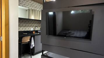 Una habitación moderna con un televisor de pantalla plana en la pared, un escritorio pequeño con una silla y una zona de cocina al fondo.