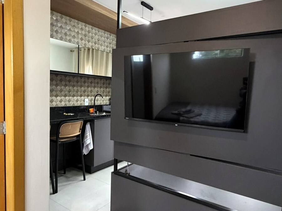 Una habitación moderna con un televisor de pantalla plana en la pared, un escritorio pequeño con una silla y una zona de cocina al fondo.