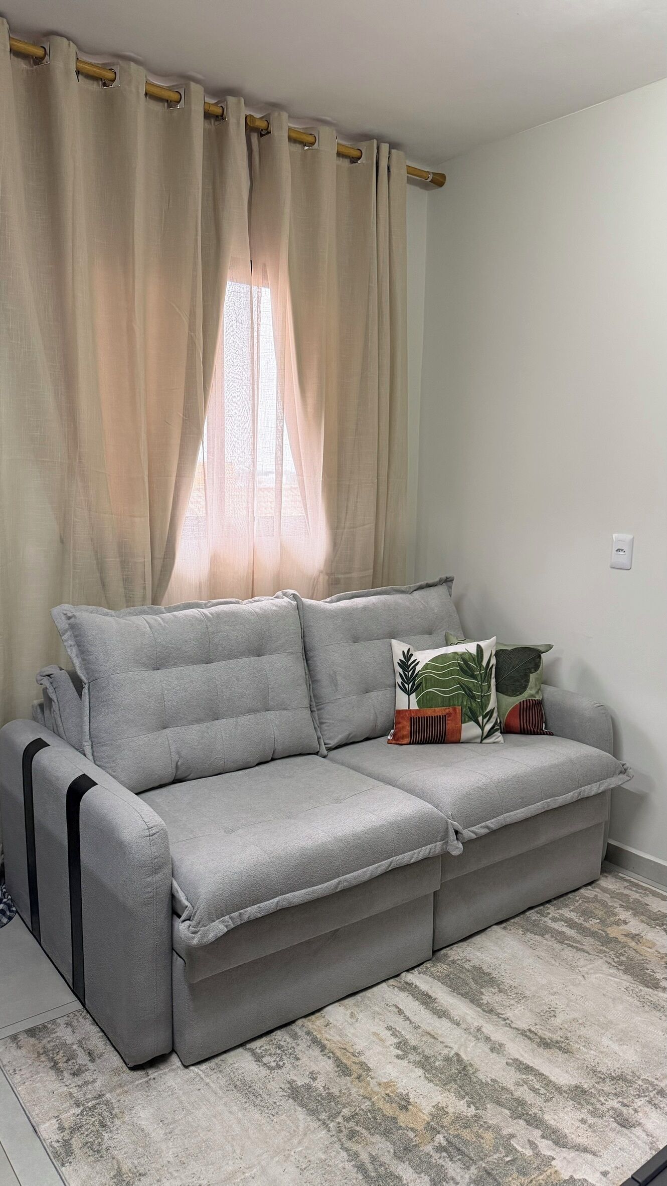 Um sofá cinzento com uma almofada, um travesseiro estampado e uma janela com cortinas.