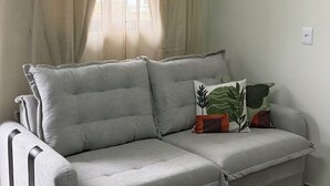 Um sofá cinzento com uma almofada, uma janela com cortinas bege e uma tomada na parede.