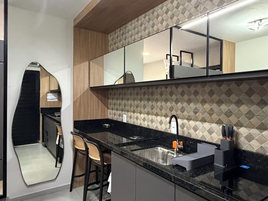 Una cocina moderna con encimera negra, un lavamanos y un espejo encima. En la zona de comedor contigua hay dos sillas y una mesa pequeña.