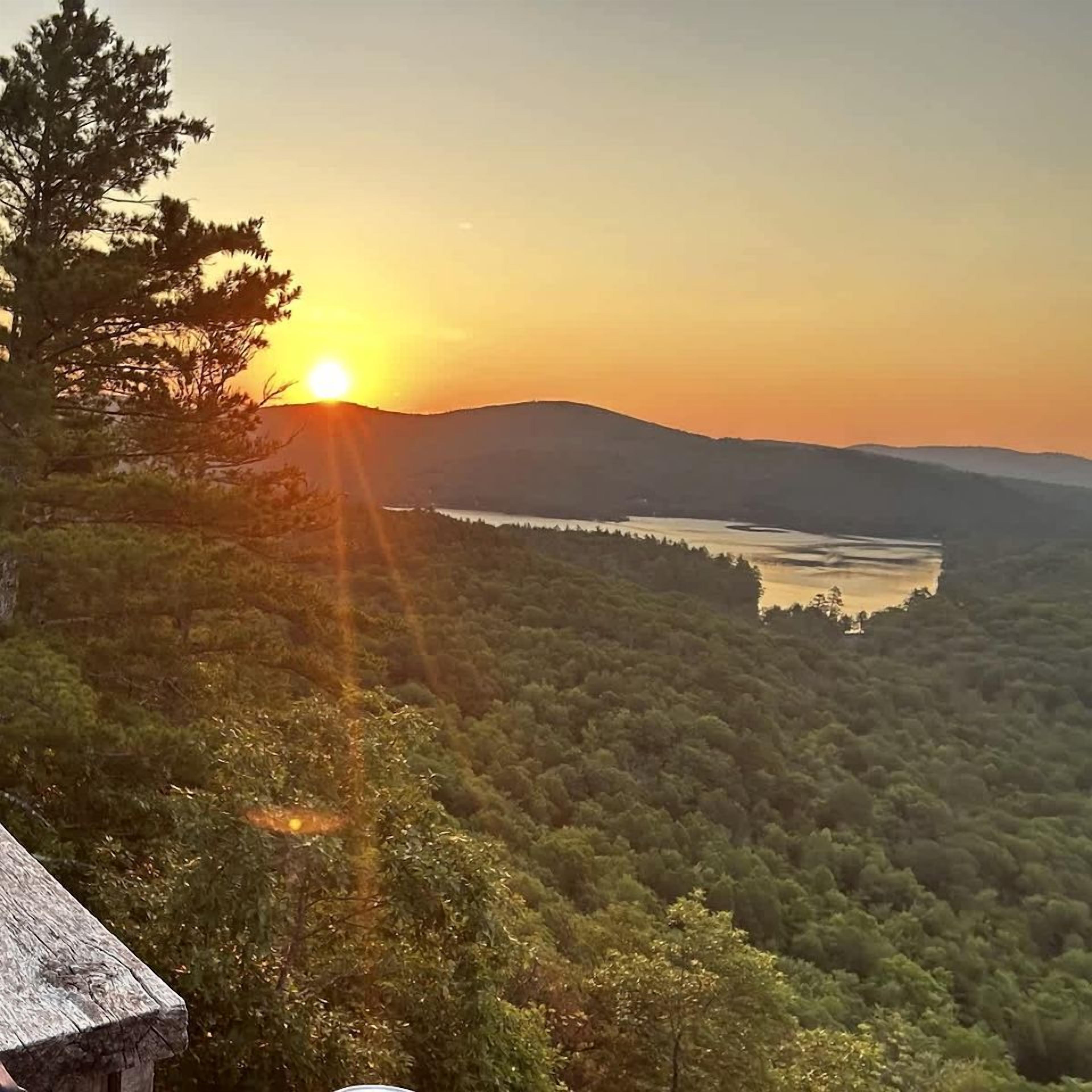 Une vue sur le coucher de soleil, avec un paysage boisé, un plan d’eau et des collines au loin.