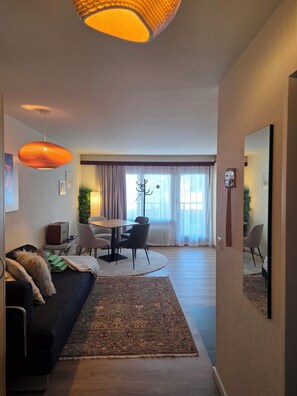 A modern living room with a sofa, a dining table with chairs, a large rug, and a ceiling light fixture.
