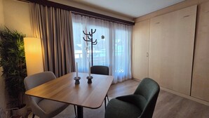 A hotel room with a wooden table, two chairs, a lamp, a coat rack, and a large window with curtains. - Cozy studio, centrally located, with mountain views (Saas-Fee)