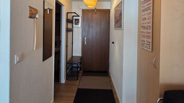 A hallway with a wooden floor, a black mat at the entrance, a hanging light fixture, and a door with a wooden frame. - Cozy studio, centrally located, with mountain views (Saas-Fee)