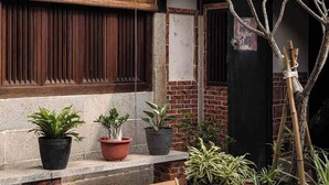 Una mesa y sillas de madera frente a un edificio con patio de baldosas, plantas en macetas y un árbol de bambú.