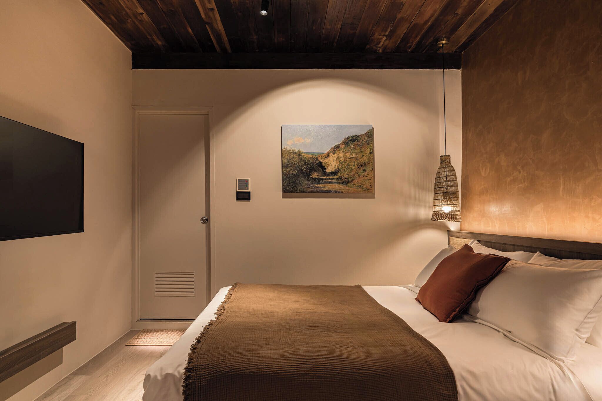 A bedroom with a bed, a painting on the wall, a door, and a hanging light fixture.