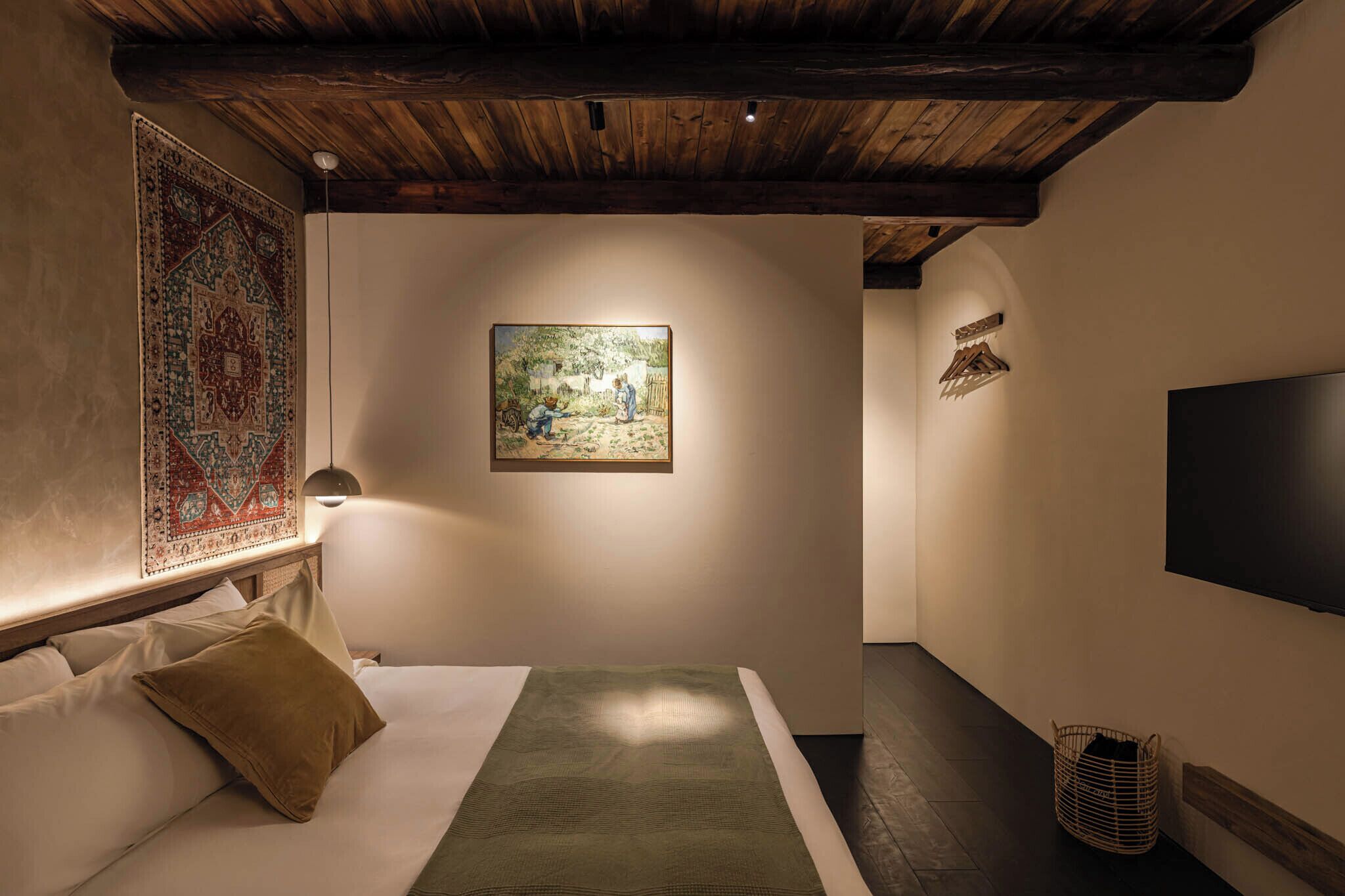 A bedroom with a bed, a hanging tapestry, a painting, a television, and a woven basket.