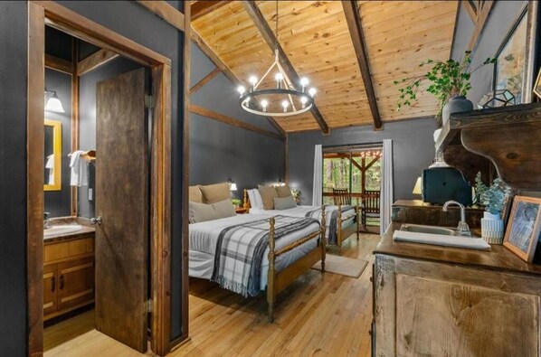 A bedroom with a wooden ceiling, a chandelier, a bed with a plaid blanket, and a wooden dresser with a sink.