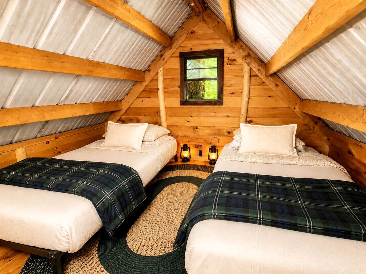 A cozy attic bedroom with two beds, a window, and a rustic wooden ceiling.